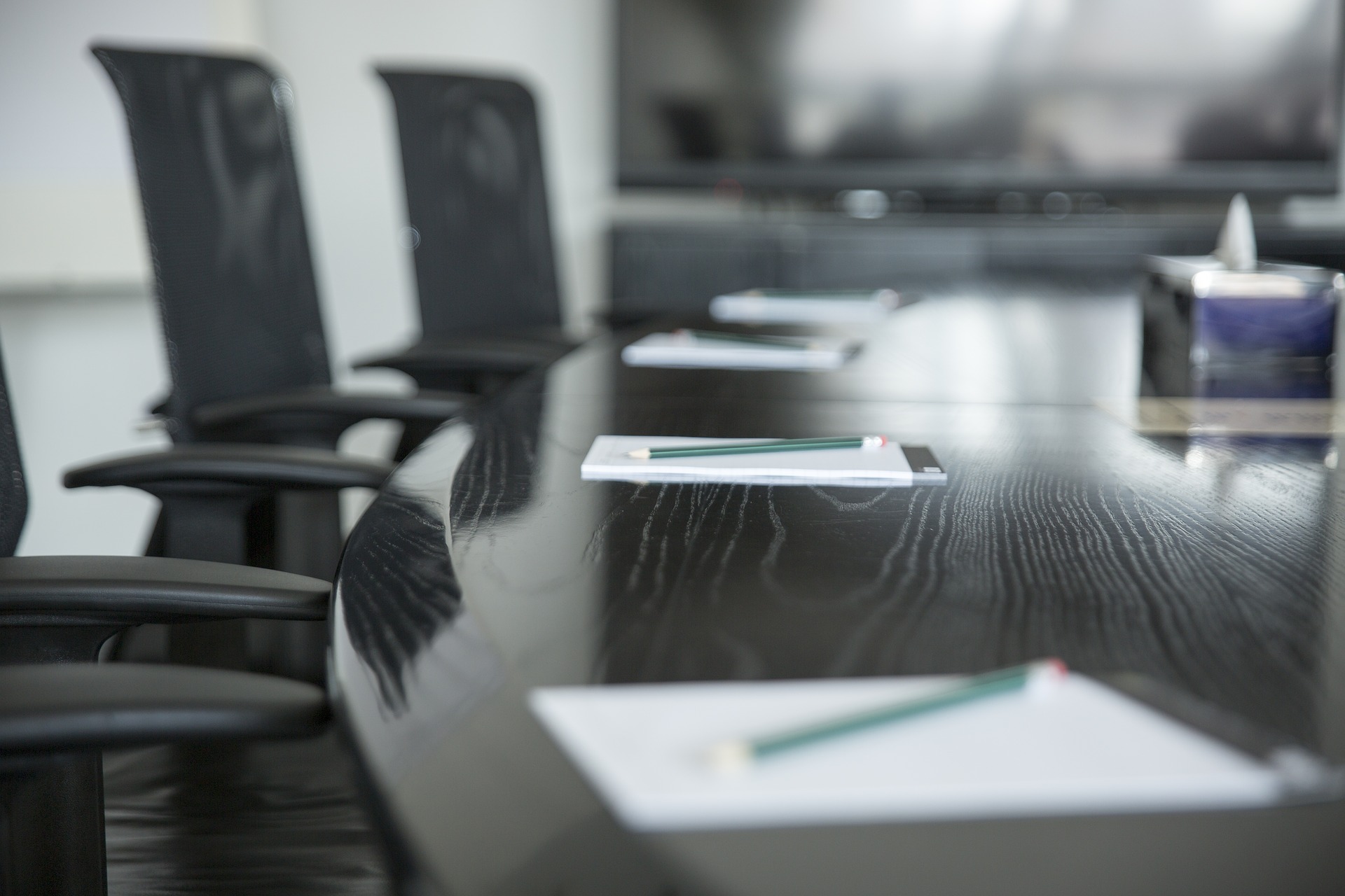 A close up of a black wood conference table with paper and pencils on it, surrounded by empty chairs.