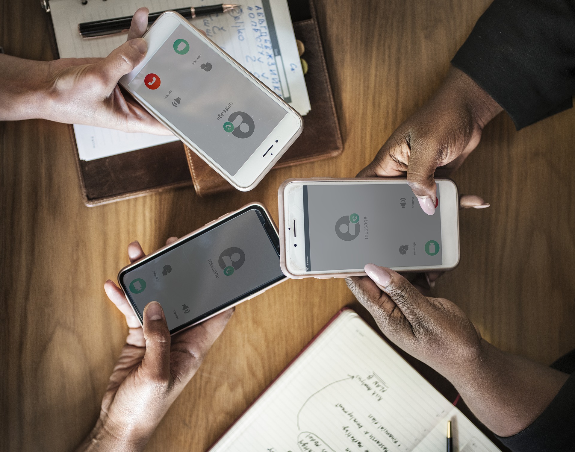 The phones are buzzing! This image has 3 people whose phone's are ringing, and they are holding them over a table with notebooks on them.